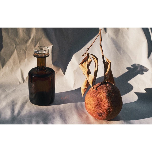 Still life featuring a brown glass bottle and a dried orange with a branch, set against a softly wrinkled white background.