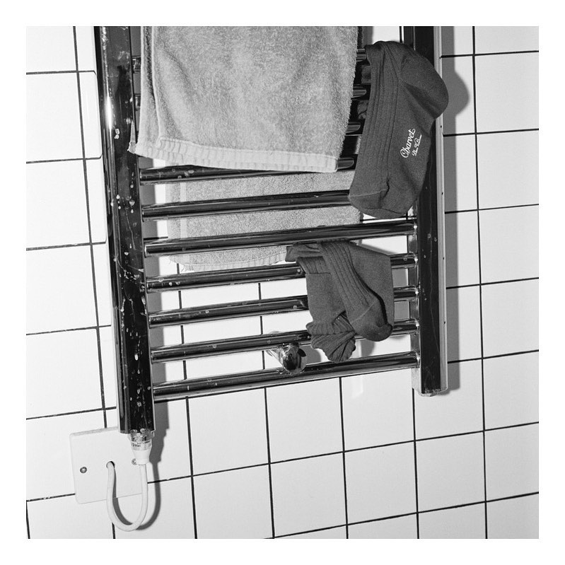 Black and white photograph of a chrome radiator with a gray towel and gray socks hanging on it in a tiled bathroom indoors.