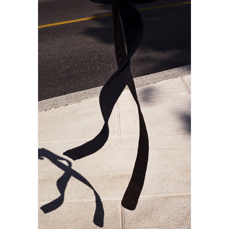 Abstract view of a black, rippled ribbon-like shape against a concrete sidewalk and asphalt roadway in full sunlight.