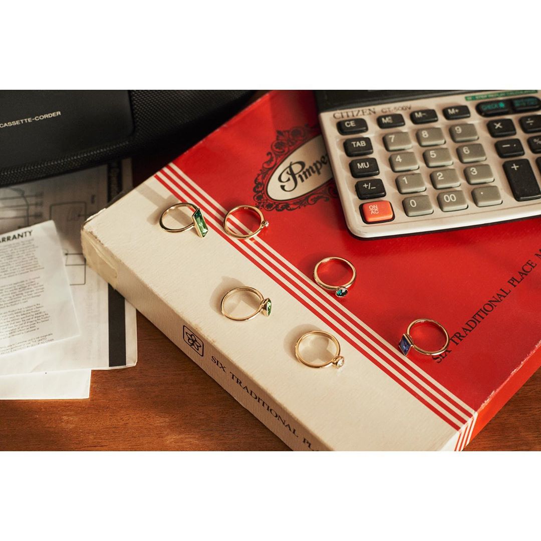 Close-up of a collection of gold rings with gemstones on a red book, alongside a vintage calculator and office accessories.
