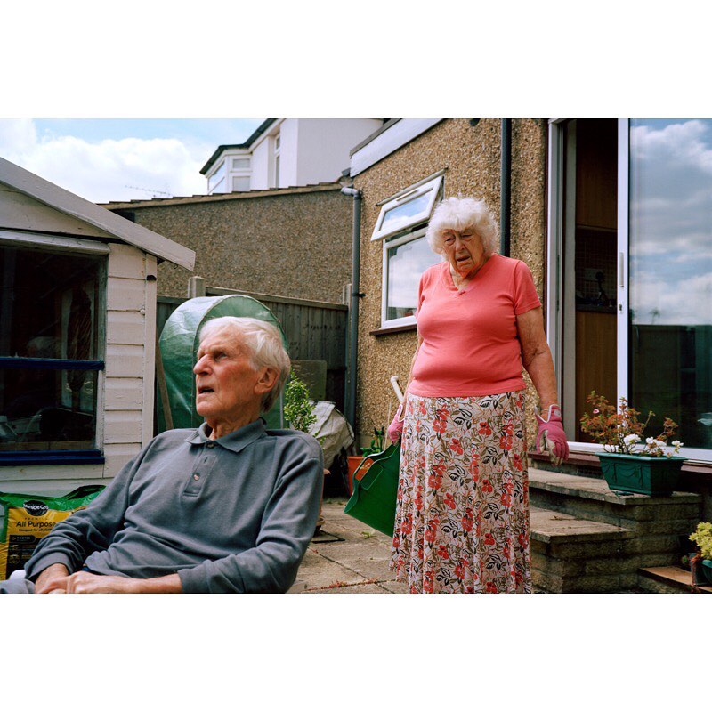 Elderly couple relaxing outdoors in garden. Woman in floral skirt stands beside seated man in grey polo shirt in backyard on sunny day.