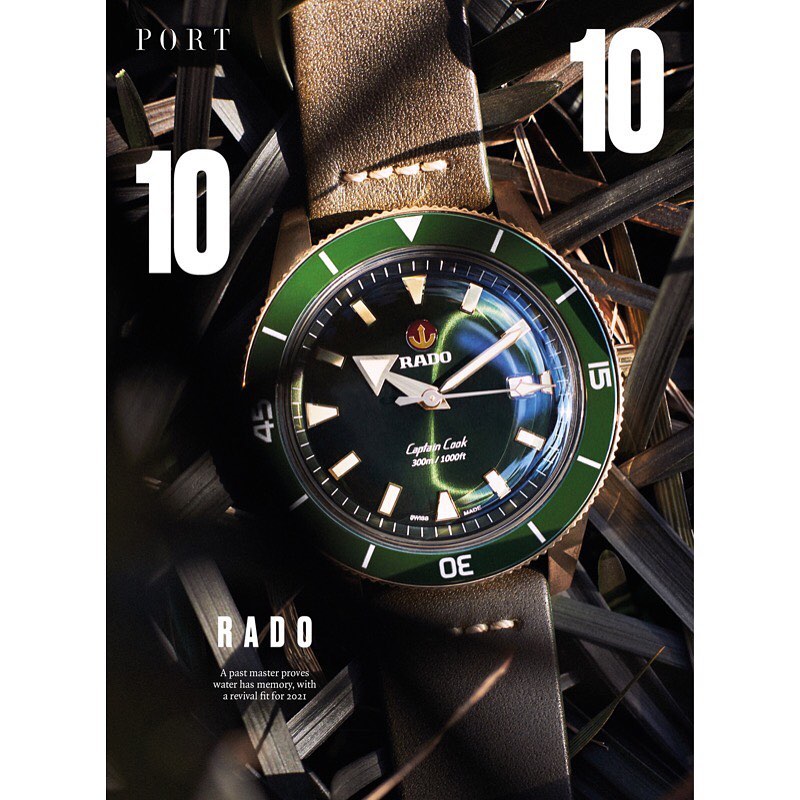 Close-up shot of a RADO Captain Cook watch on a grey-brown leather strap, surrounded by dark green foliage, in a studio setting.