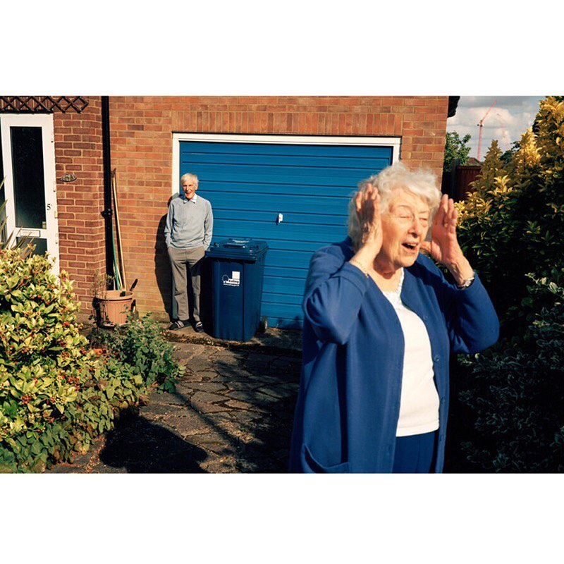 Older woman reacting with hands on head in yard; man in background. Blue garage door, brick house, and greenery visible in outdoor scene.