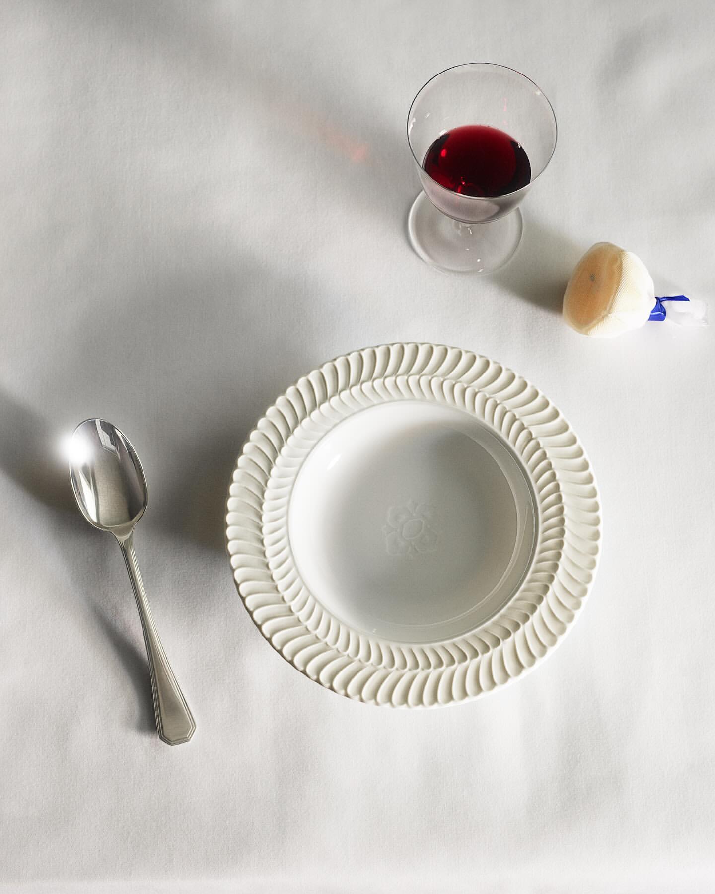 Overhead shot of a white decorative plate, silver spoon, glass of red liquid, and cleaning putty on a white surface, food and beverage