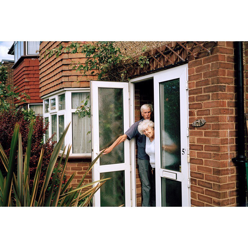 Elderly couple standing in the doorway of a brick house with green plants, wearing casual clothing, in daylight.