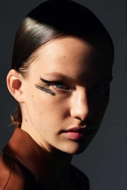 Close-up portrait of a young woman with slicked-back dark hair and thick black markings above her eye, wearing a brown jacket.
