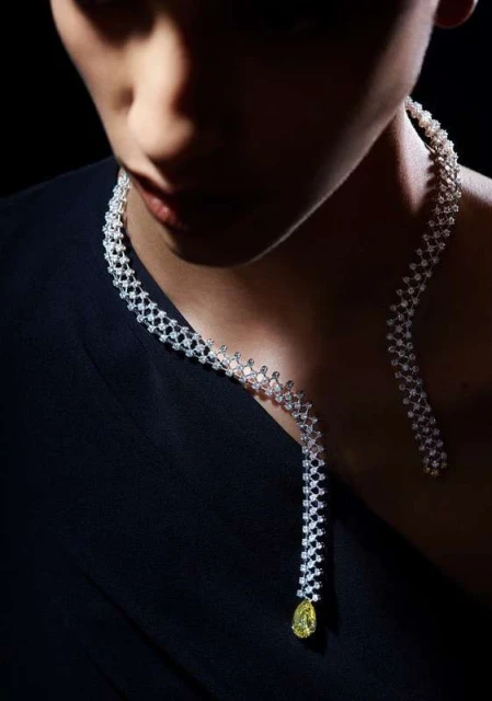 Elegant woman wearing a sparkling diamond necklace with yellow pendant on black draped fabric against a dark, blurred background.
