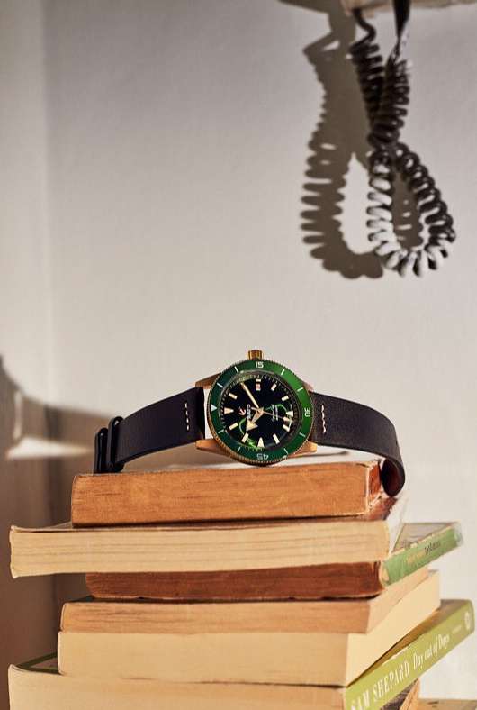 Elegant wristwatch with a green bezel and a black leather strap sits on a stack of antique books against a cream-colored wall.