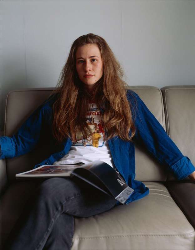 Young adult female sitting on a beige sofa, holding an open book. She is wearing a blue cardigan and a white t-shirt with a graphic.
