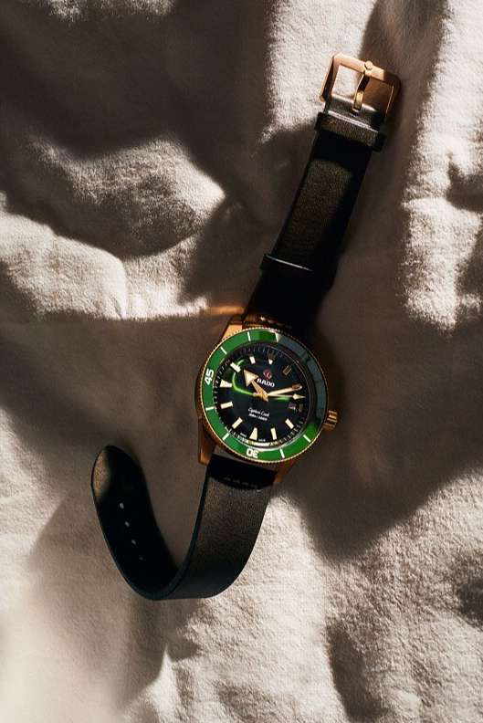 Wristwatch close-up on textured white fabric background. Gold-tone watch casing, dark green dial, black leather strap visible.