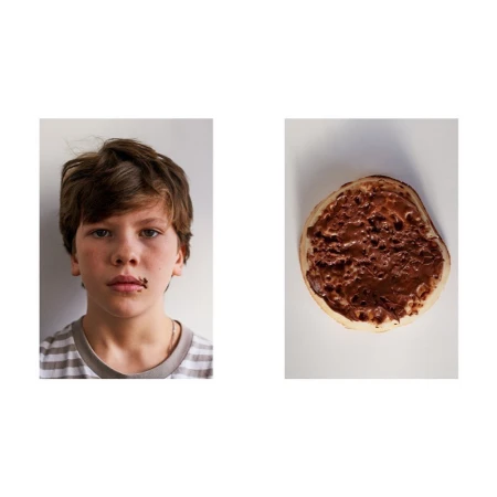 A young person with brown hair is seen with chocolate around their mouth, next to a pancake covered in chocolate spread on a white background.