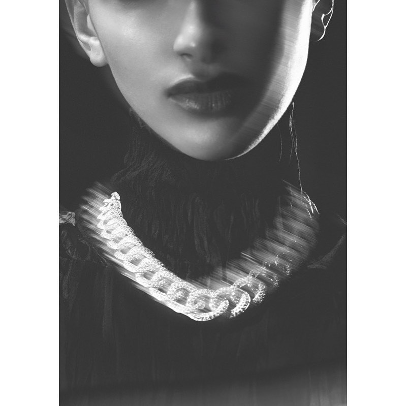 Monochrome portrait of a person with dark hair and lipstick wearing a chain necklace, set against a dark background.