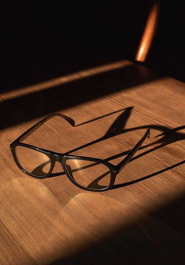 A pair of black-rimmed eyeglasses sit on a wooden surface with light casting shadows across the frame in a warm, inviting indoor setting.