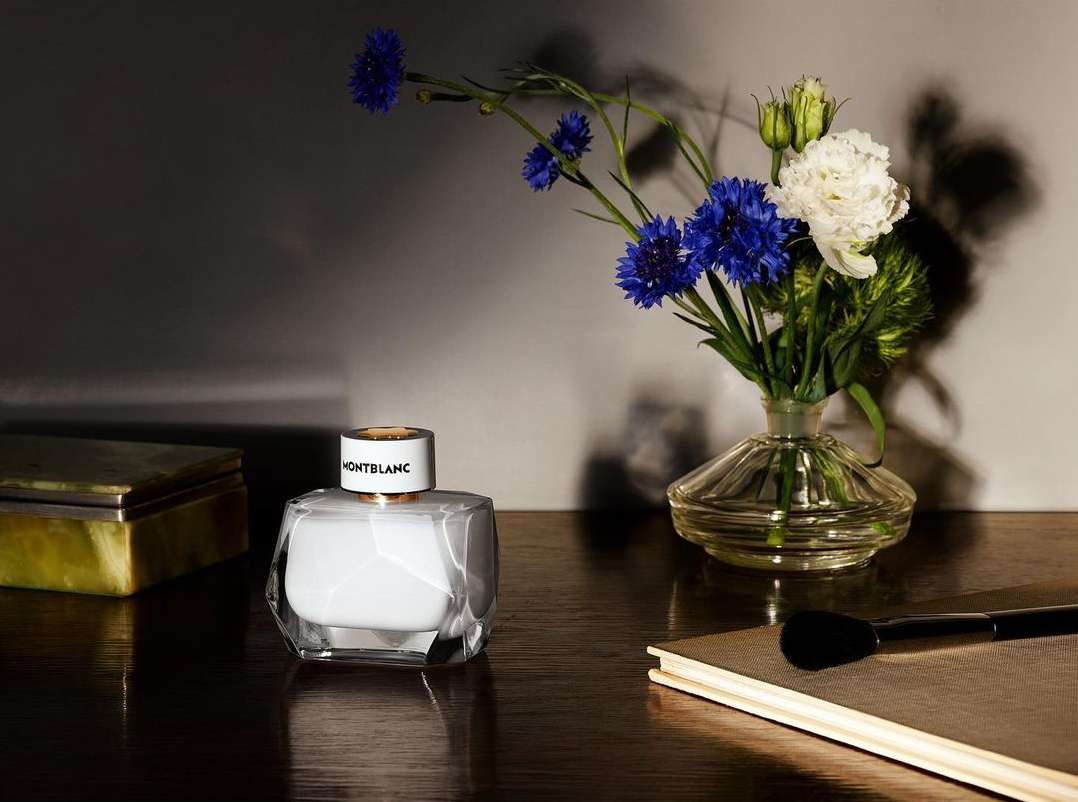 Product still life featuring a Montblanc perfume bottle alongside flowers in a vase, a box, and a makeup brush on a wooden surface.