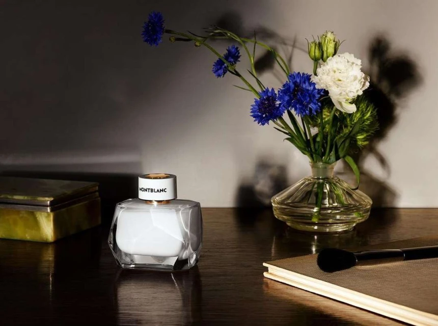 Product still life featuring a Montblanc perfume bottle alongside flowers in a vase, a box, and a makeup brush on a wooden surface.