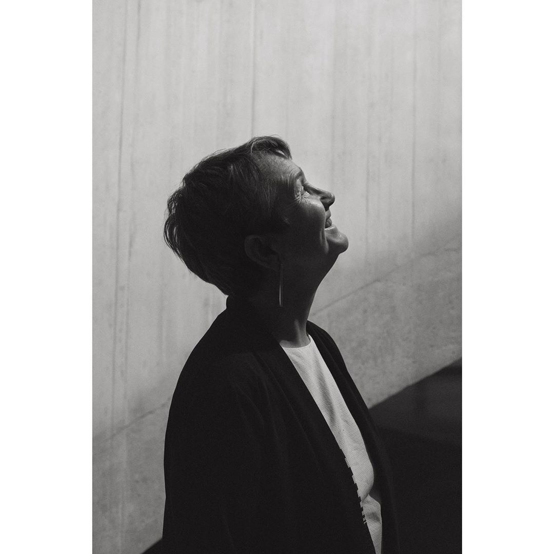 Profile portrait of a smiling older woman with short hair looking up, wearing a dark jacket against a gray concrete background.