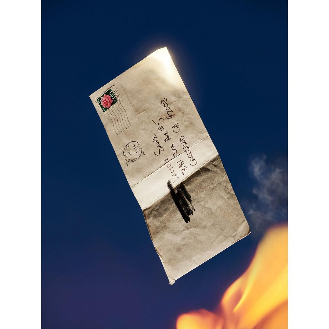 A burnt envelope with handwritten address floats in a deep blue sky with bright flames at the bottom, creating a dramatic visual.