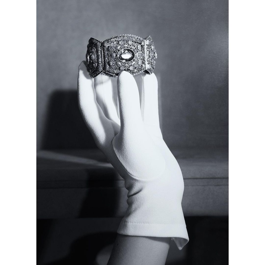 Close-up of a gloved hand holding a vintage ornate silver bracelet, against a soft grey backdrop, emphasizing details and elegance.