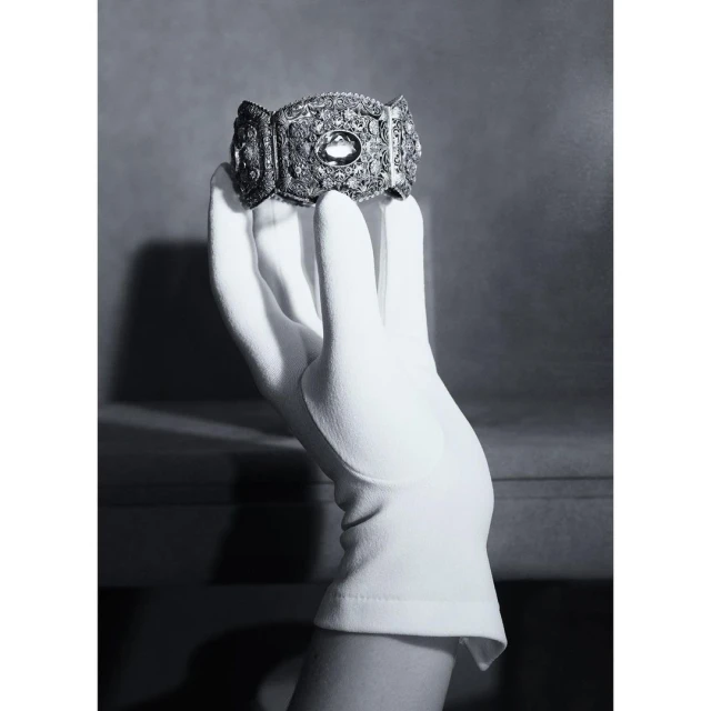 Close-up of a gloved hand holding a vintage ornate silver bracelet, against a soft grey backdrop, emphasizing details and elegance.