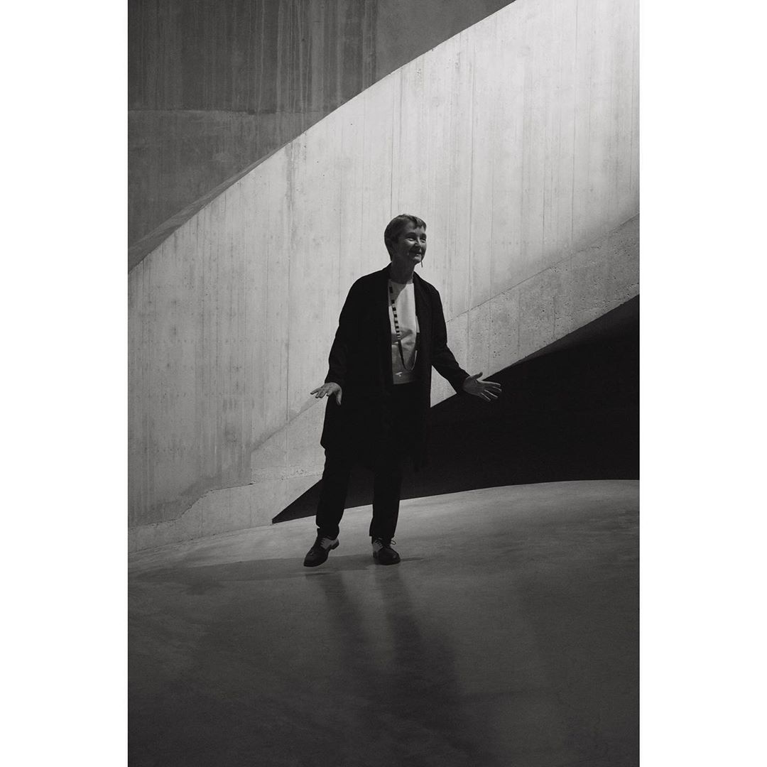 Black and white portrait of an adult woman standing indoors in front of a concrete wall and staircase in a wide shot.