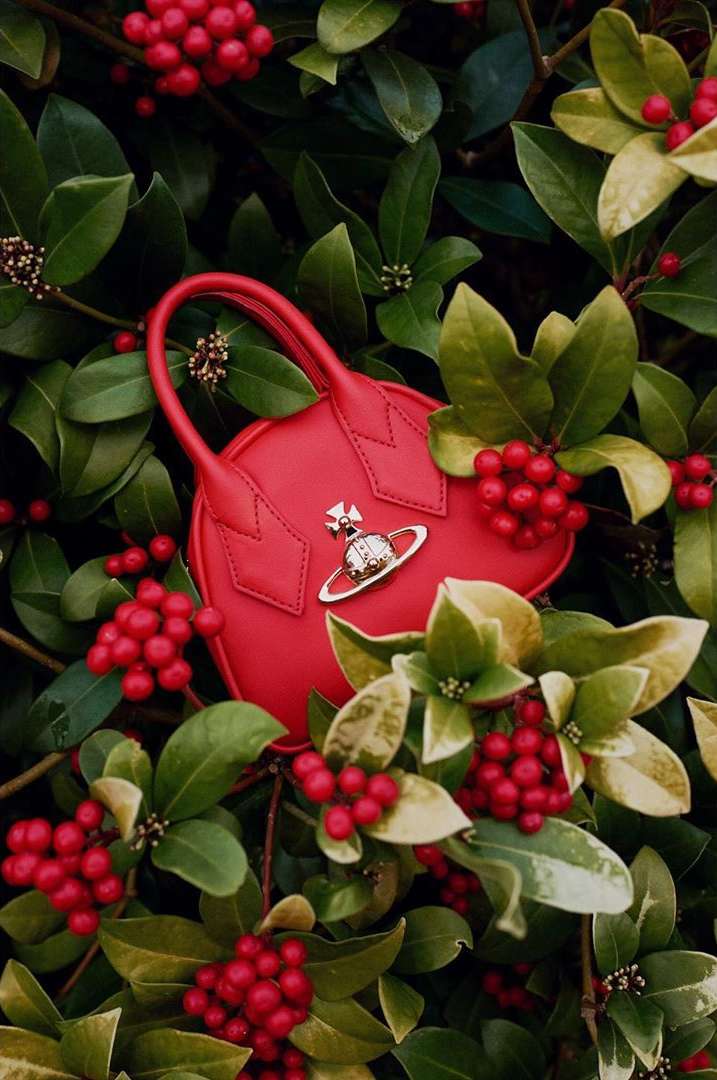 Red Vivienne Westwood handbag with orb logo on a bush with red berries and green leaves, fashion accessory still life.