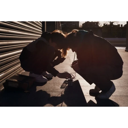Two people squatting outdoors, focusing on connecting electronic components on the ground, with a box, building facade, and roller door.