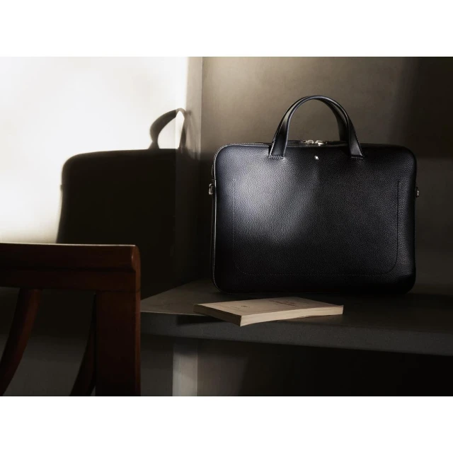 Leather briefcase with a handle and a book are staged indoors with natural light and shadow creating a sophisticated, professional setting.