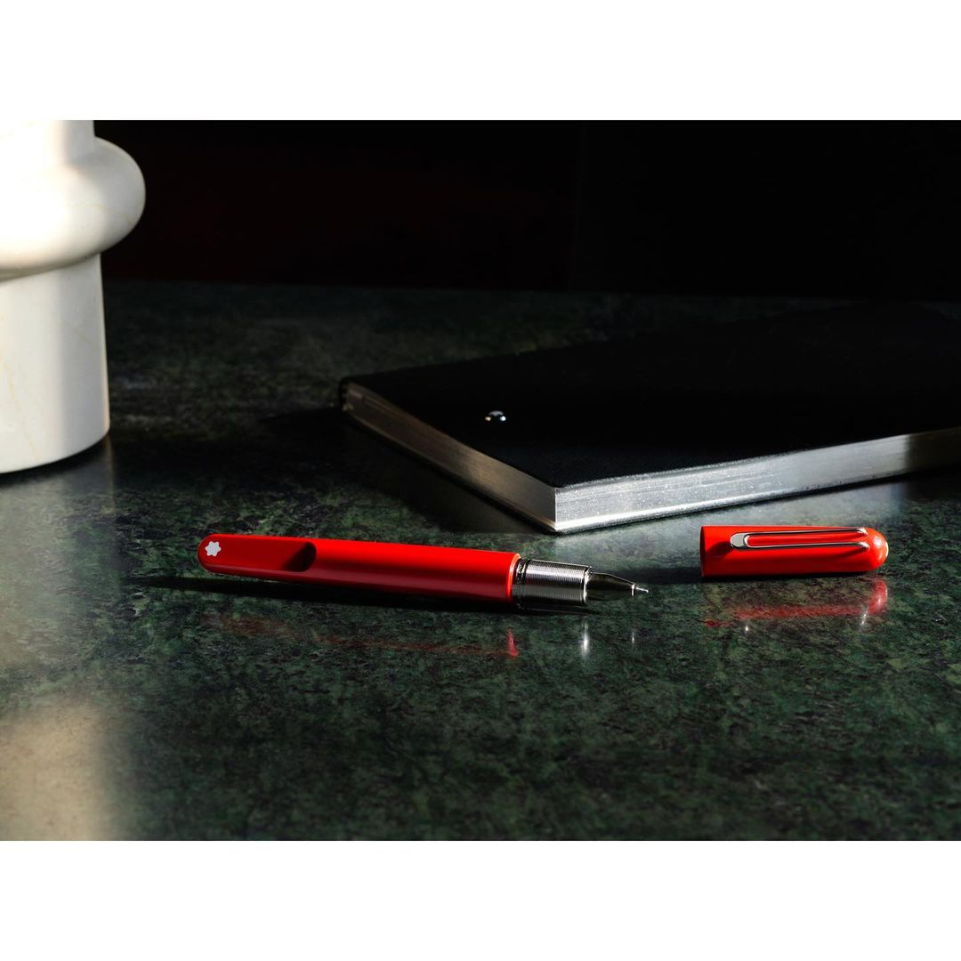 A red fountain pen and its cap are laid on a dark green marble surface, with a black notebook and white vase in the background.