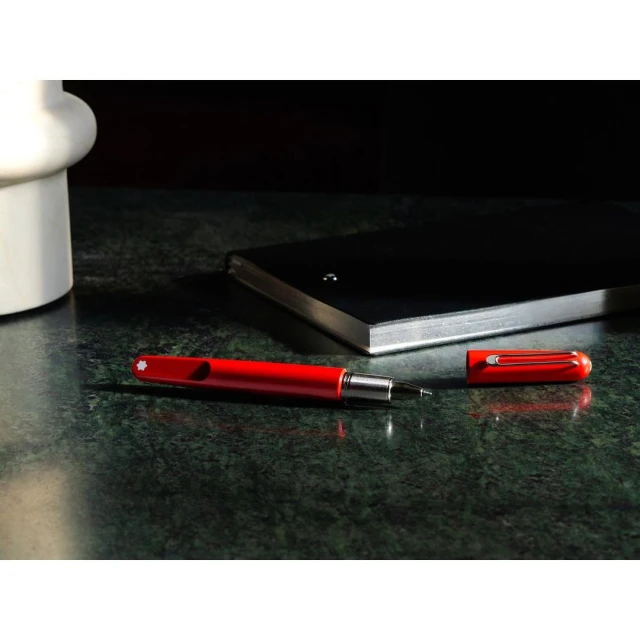 A red fountain pen and its cap are laid on a dark green marble surface, with a black notebook and white vase in the background.