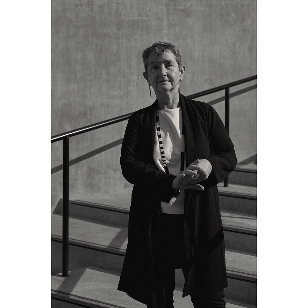 Black and white portrait of a mature woman with short hair, wearing a dark coat and standing on concrete steps with a metal railing.