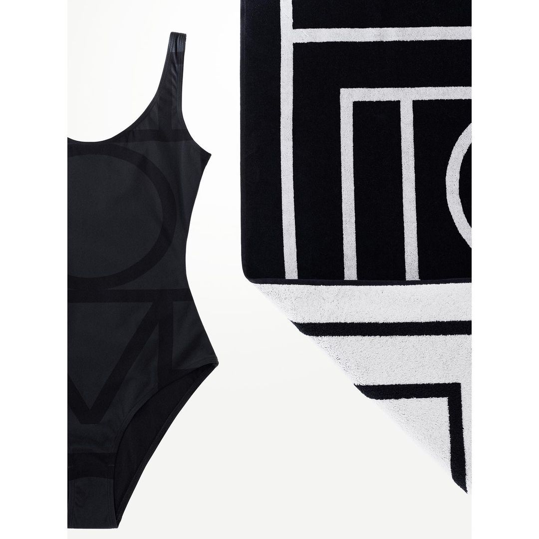 Black one-piece swimsuit with oversized LOVE text and a black and white geometric towel on a white background. Minimalist swim fashion.