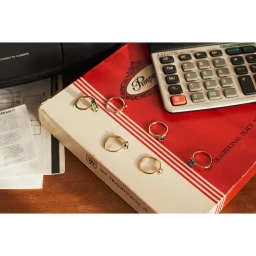 Several gold rings with colorful gemstones arranged diagonally on a red ledger book alongside a calculator.
