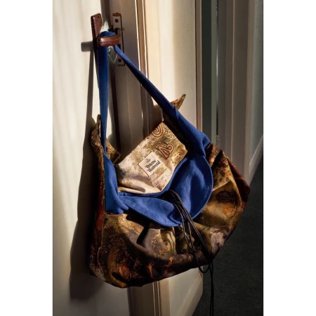 A patterned fabric shoulder bag with blue straps hanging on a wooden wall hook in interior light.