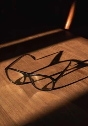 Black eyeglasses resting on a warm wooden tabletop under dramatic directional lighting casting sharp shadows.