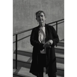 Monochrome portrait of an older woman standing on outdoor steps by a metal railing, captured by Tex Bishop.