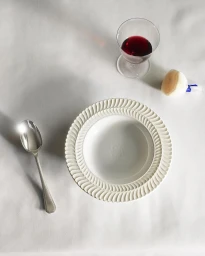 An overhead still life composition with a white decorative plate, a silver spoon, a glass of red wine, and a small brush on a white tablecloth.