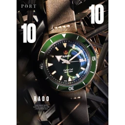 Close-up of a Rado wristwatch with a green bezel and brown leather strap resting among dark green grass blades.
