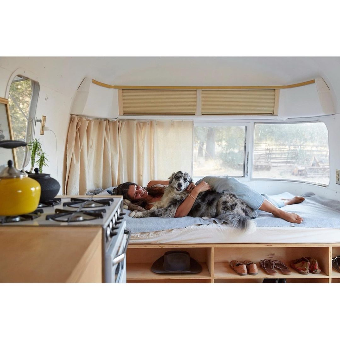 A woman and her dog are relaxing on a bed inside an Airstream trailer, with a view of the outdoors through a large window, during daytime.