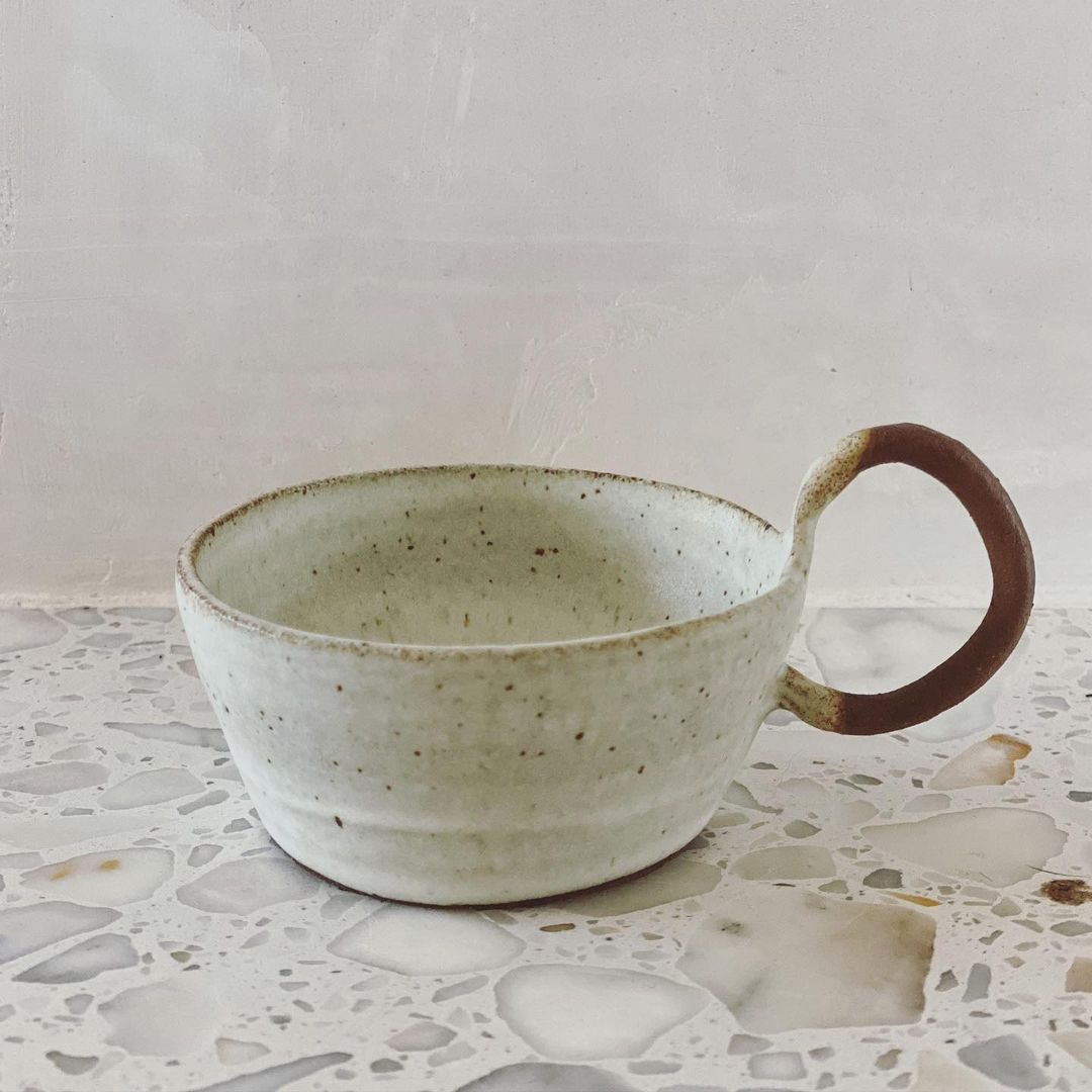 Close-up of a speckled ceramic mug with an exaggerated handle, sitting on a terrazzo countertop in a softly lit indoor setting.