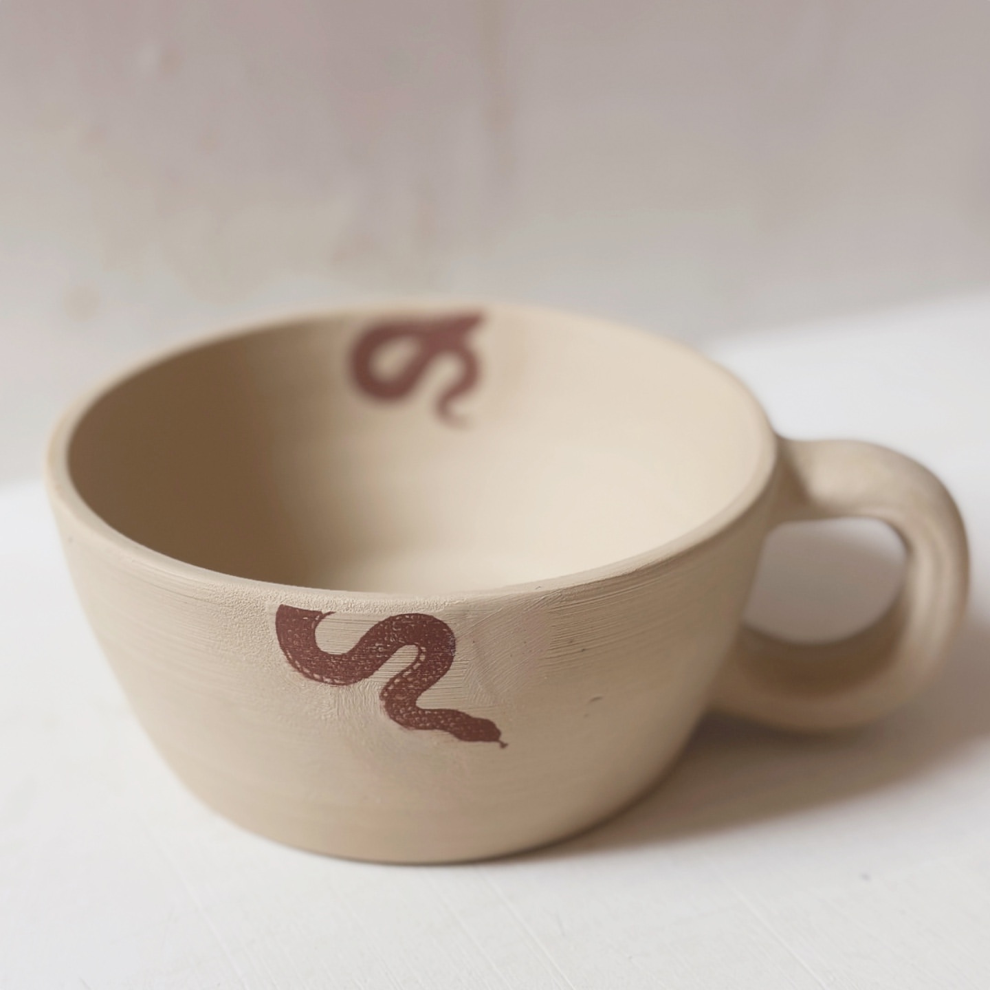 Beige ceramic cup with handle, adorned with dark brown snake pattern, clean minimal background, studio lighting, neutral aesthetic.