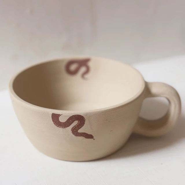 Beige ceramic cup with handle, adorned with dark brown snake pattern, clean minimal background, studio lighting, neutral aesthetic.