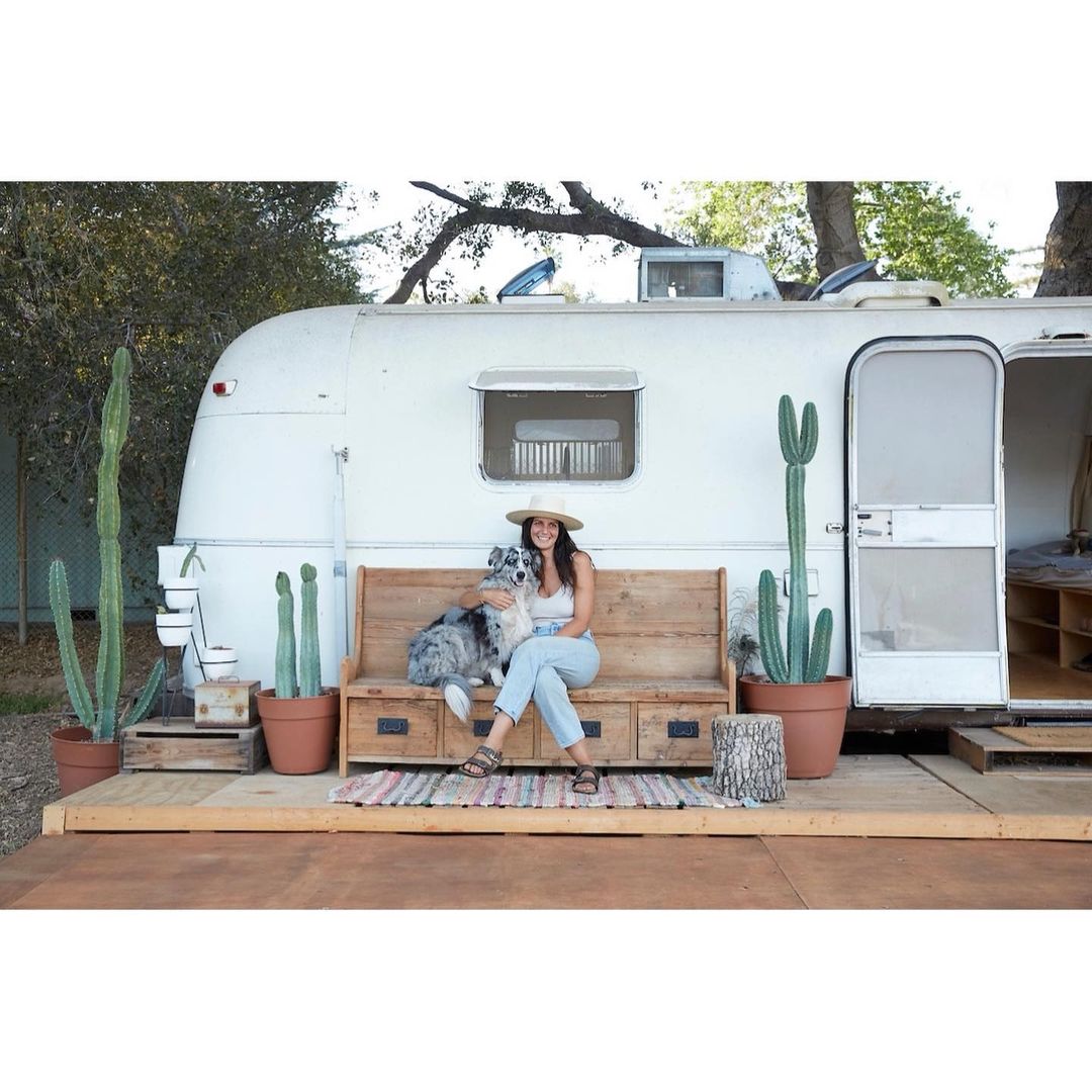 Woman sits on a bench outside an Airstream with her dog, cactus plants, and a wood deck visible. Wearing a white top and light jeans.