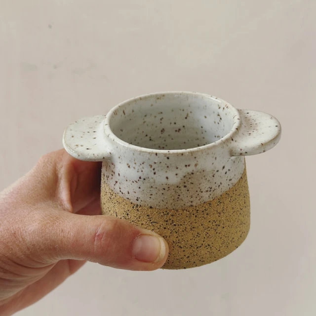 Close-up of a hand holding a two-toned ceramic mug with rounded handles against a plain background, showcasing its textured design.