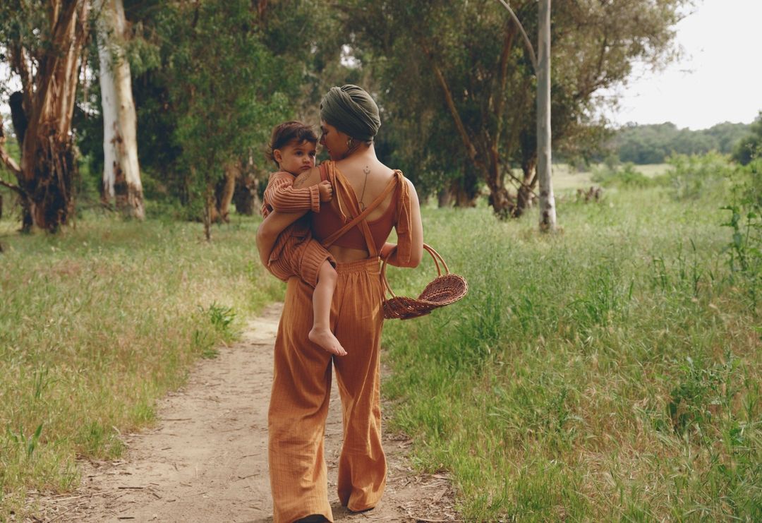 A mother carries her child on her back while walking on a dirt path through a grassy field with tall trees, carrying a woven basket.