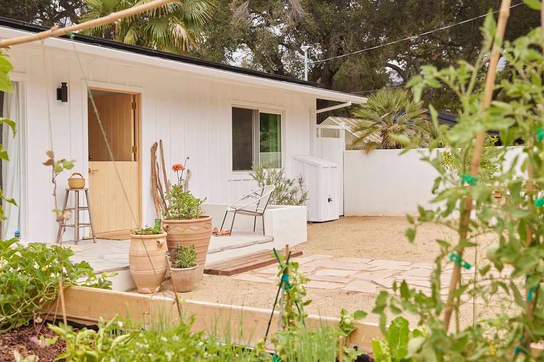 Exterior view of a white modern house with a well-maintained garden in the foreground, featuring potted plants on the patio.