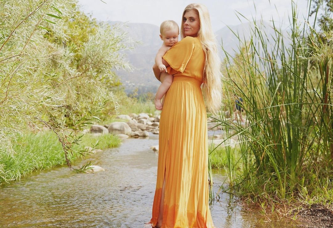 A woman in a golden dress standing in a shallow stream, holding a baby, with a backdrop of verdant trees and a gentle mountain range.
