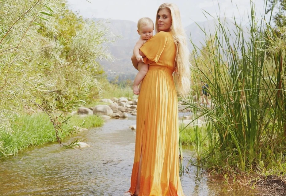 A woman in a golden dress standing in a shallow stream, holding a baby, with a backdrop of verdant trees and a gentle mountain range.