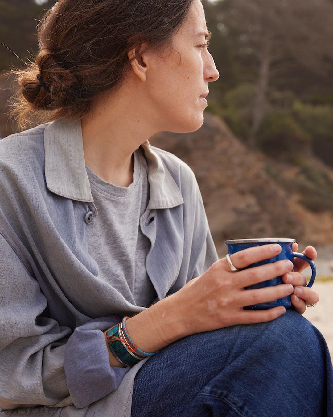 Adult woman with dark hair sits outdoors on beach holding a blue mug, wearing jeans and a button-down shirt, with blurred background.