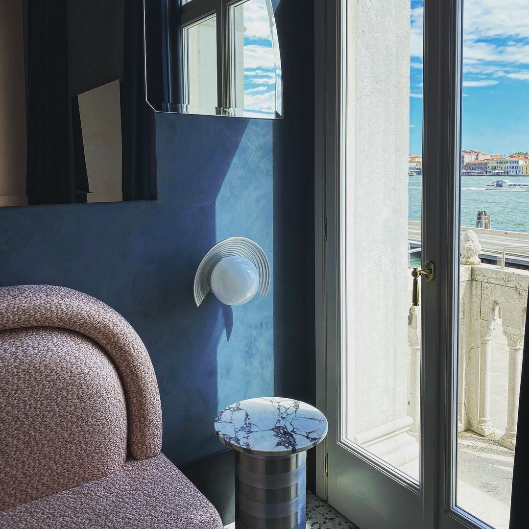 Interior of a room with a view from a window overlooking a waterway with buildings in the distance, featuring a chair and side table.