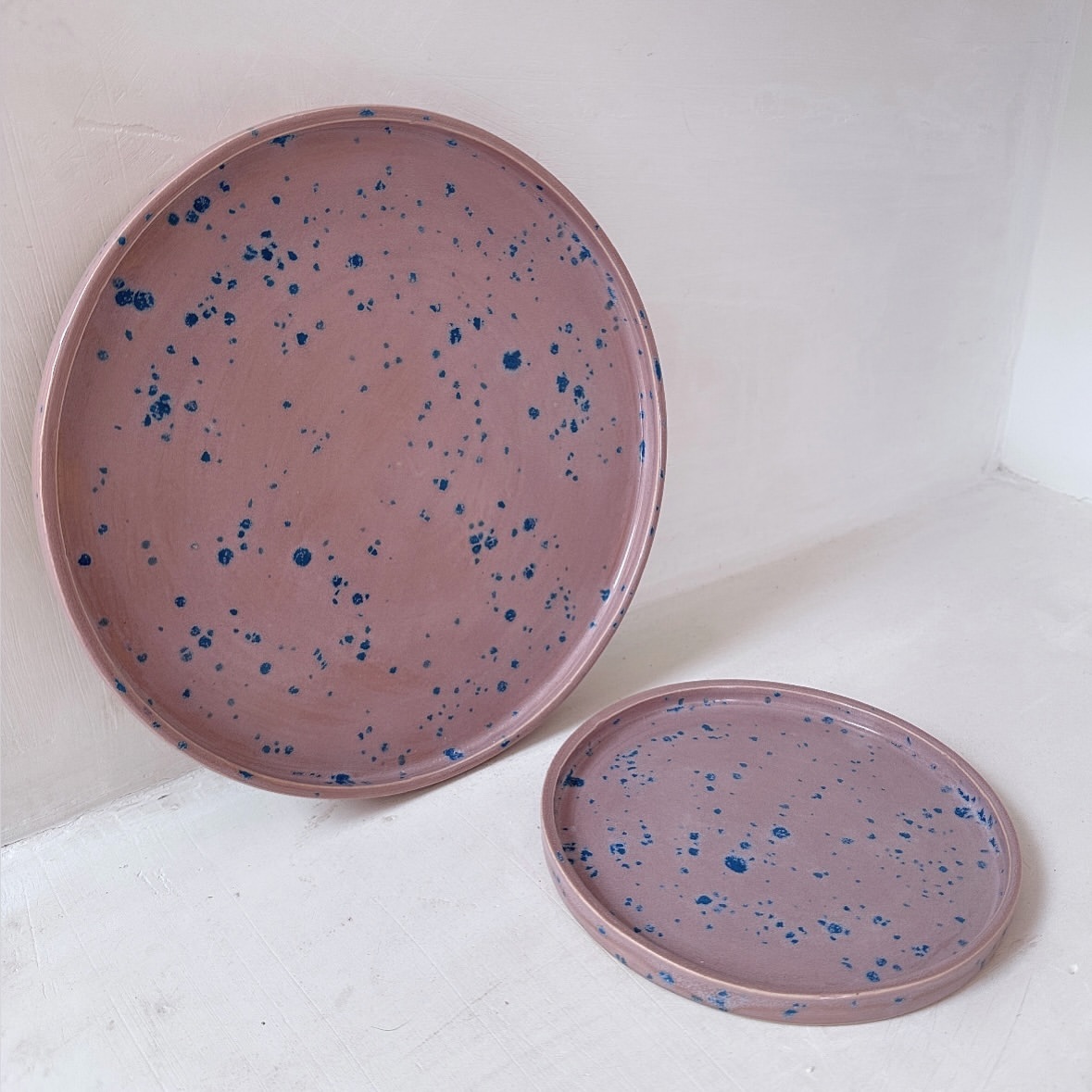 Two pink ceramic plates with blue speckles sit on a white surface, one large and one smaller, creating a minimalist, decorative scene.